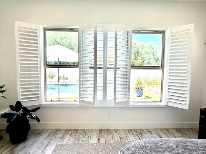 Bay window plantation shutters folded open across a bedroom wall, replacing builder blinds in a Mesa, AZ home